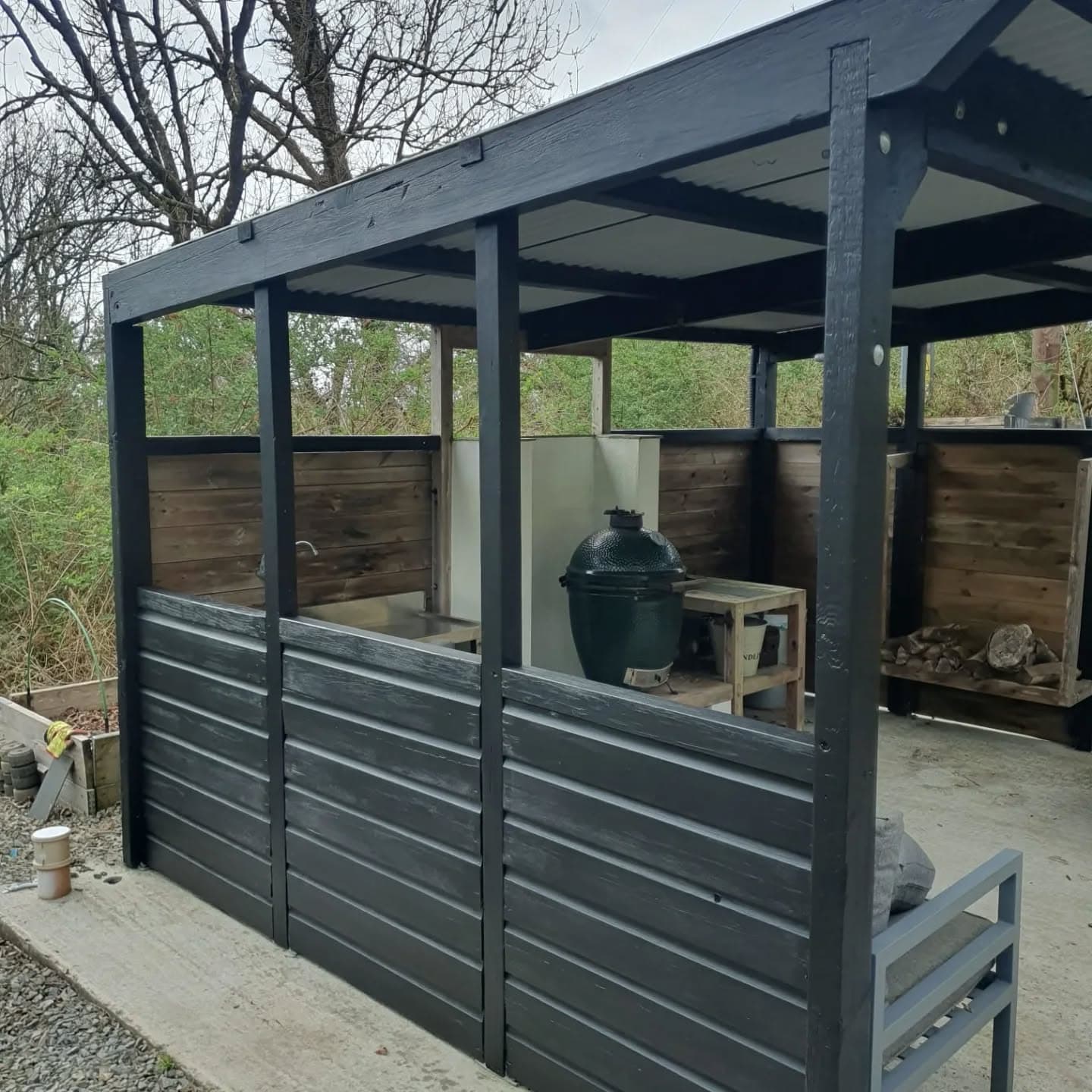 Black-painted timber outdoor BBQ pergola with shelter roof and wood store.